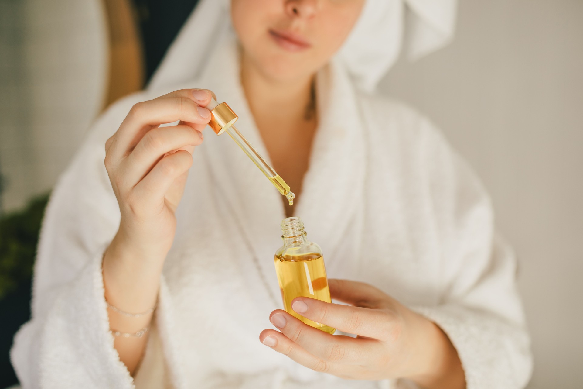 Young woman wearing white bathrobe and towel on head holding face oil in bathroom. Beauty cosmetic concept.